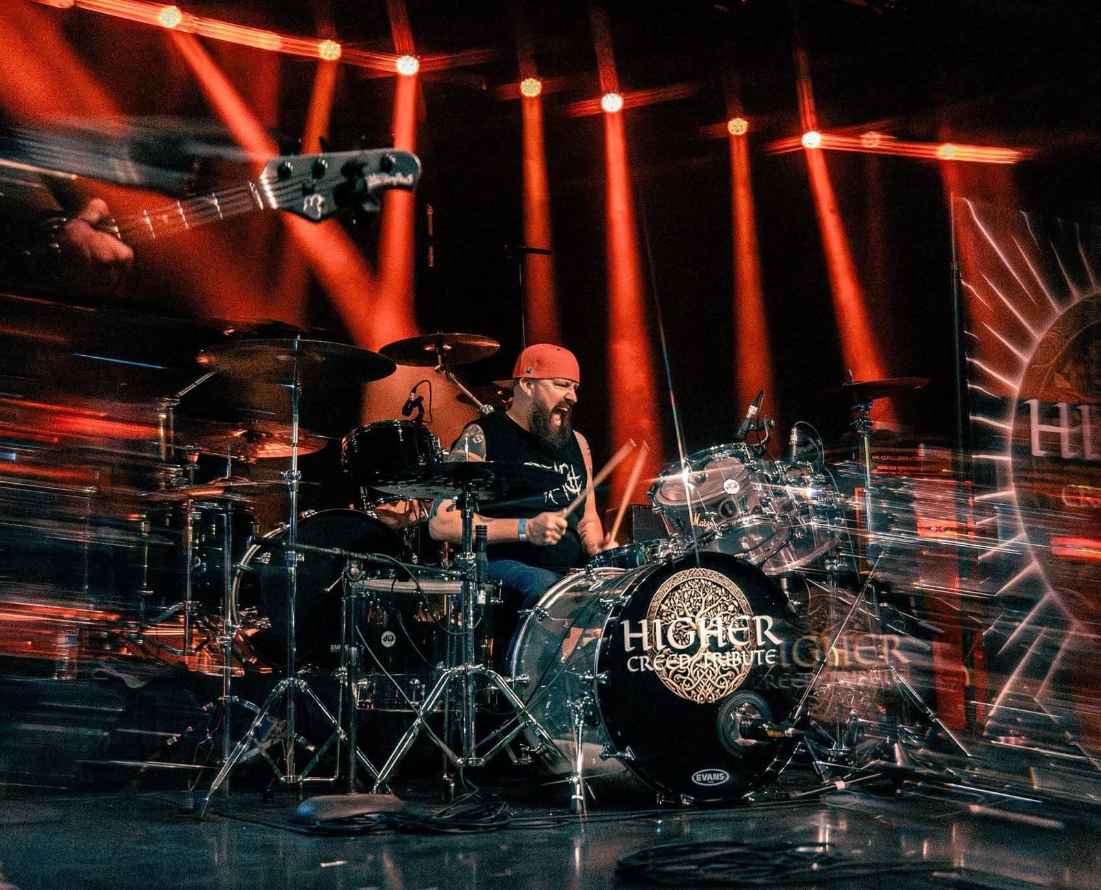Chad Edwards playing drums under red stage lights, the Higher Creed Tribute logo on his kick drum