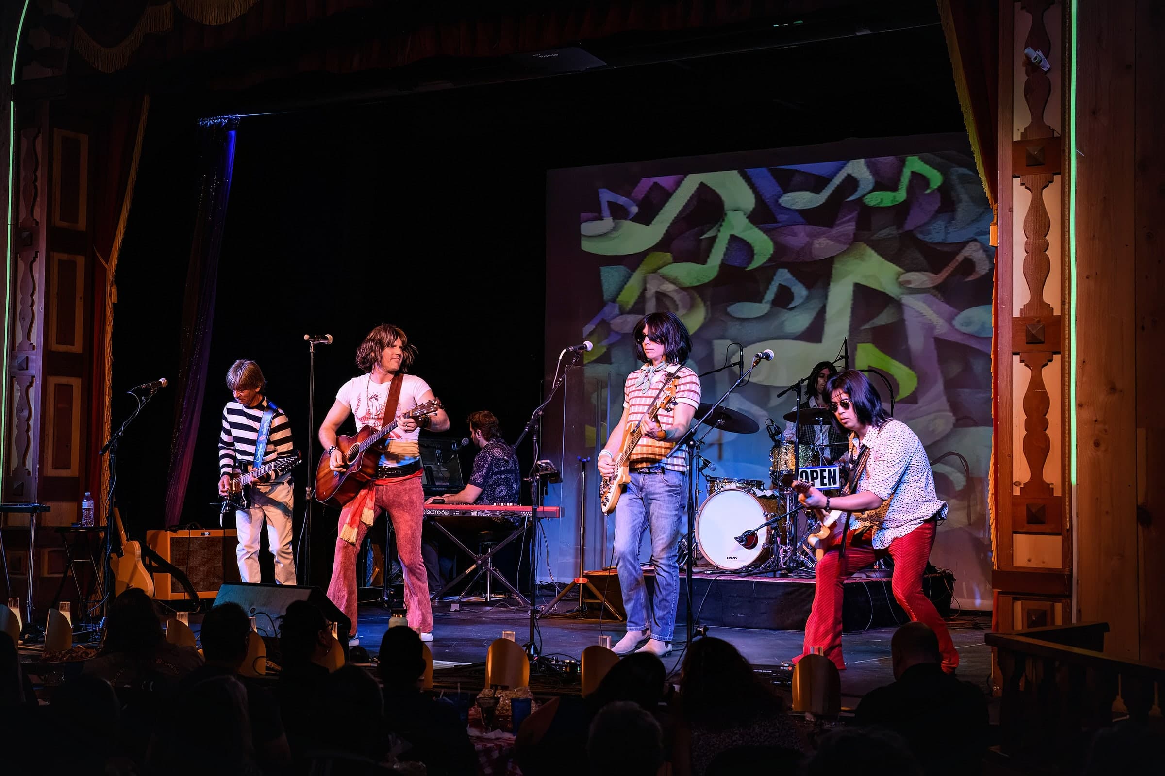 The band performing on a theater stage with a colorful projected backdrop: 2 guitarists, the singer with an acoustic guitar, a keyboard player, the bass player and the drummer, with the audience seated at tables in front