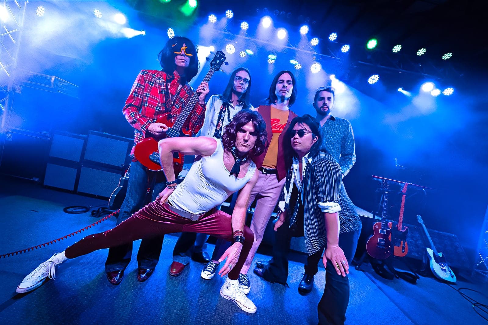 The 6 players posing on a stage under blue lights and haze, the singer in a white tank top and red velvet pants lunging in front, the bass player in a red plaid shirt behind him
