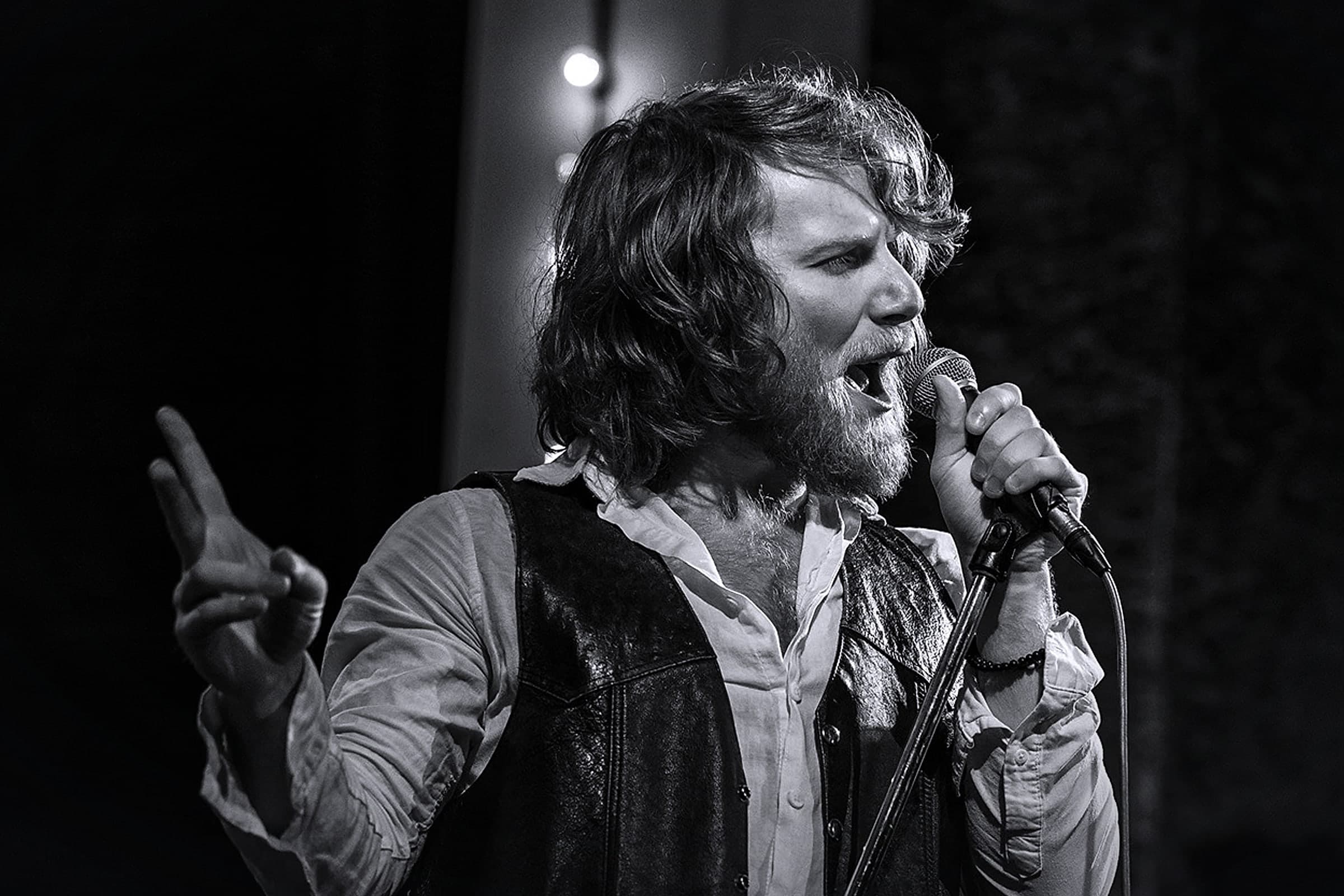 Black and white photo of the singer on stage in a leather vest and white shirt, singing into a microphone on a stand with one hand raised