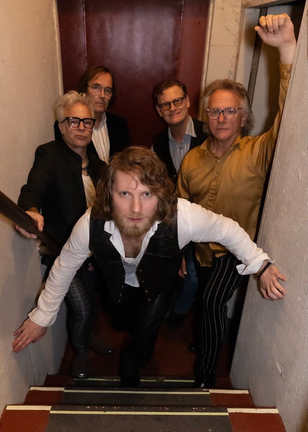 The 5 players posing in a narrow stairwell, the singer in a white shirt and vest leaning toward the camera in front and the other 4 players behind him